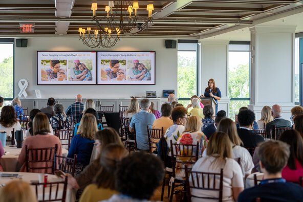 A woman at a podium speaks to a room full of people with a slide about cancer caregiving on a screen next to her.