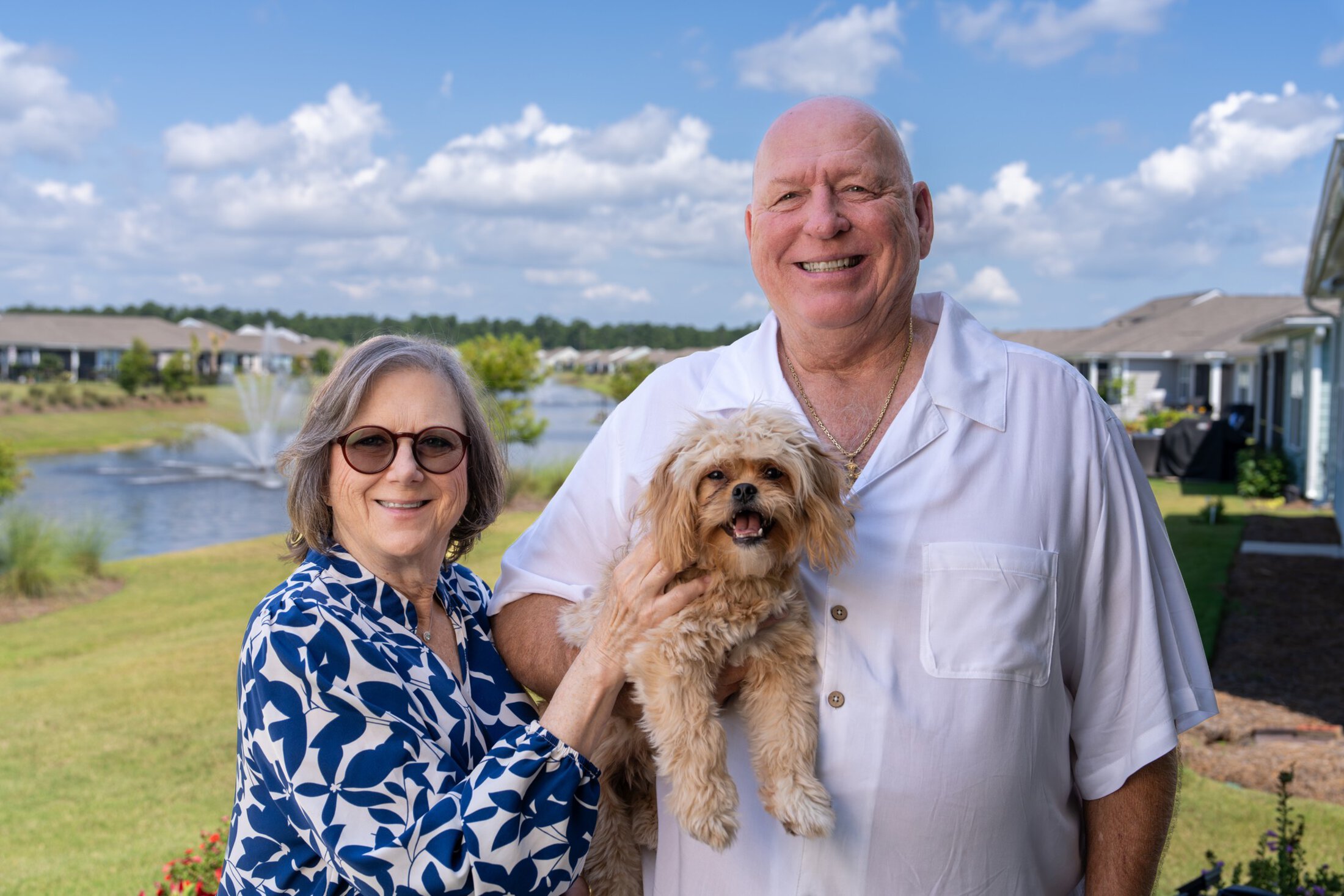 A woman and man stand together in their backyard with their small dog held between them.
