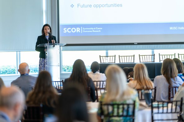 A woman speaks at a podium with many people seated at tables in front of her.