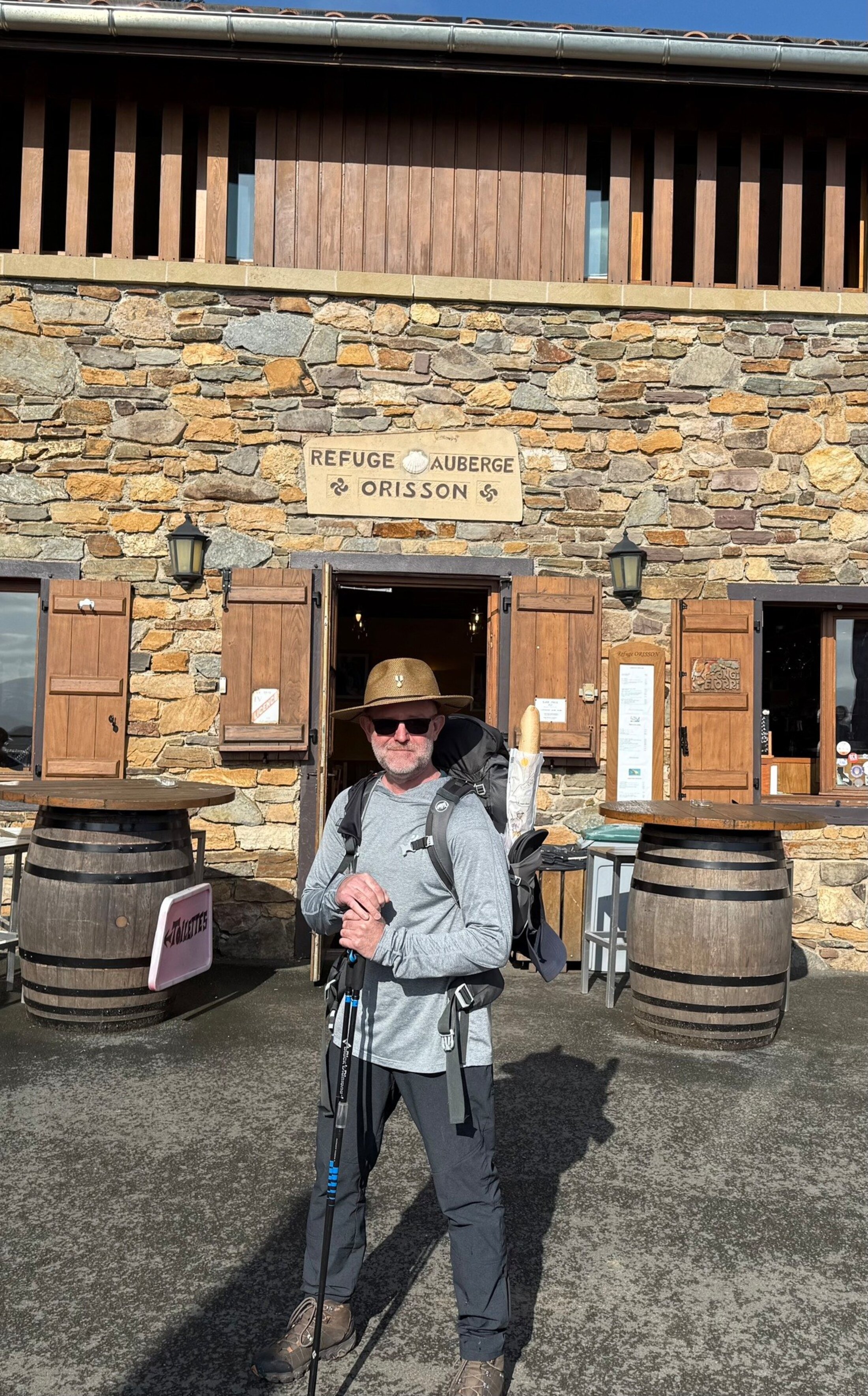 A man in hiking gear poses in front of a stone building with a sign Refuge Auberge Orisson