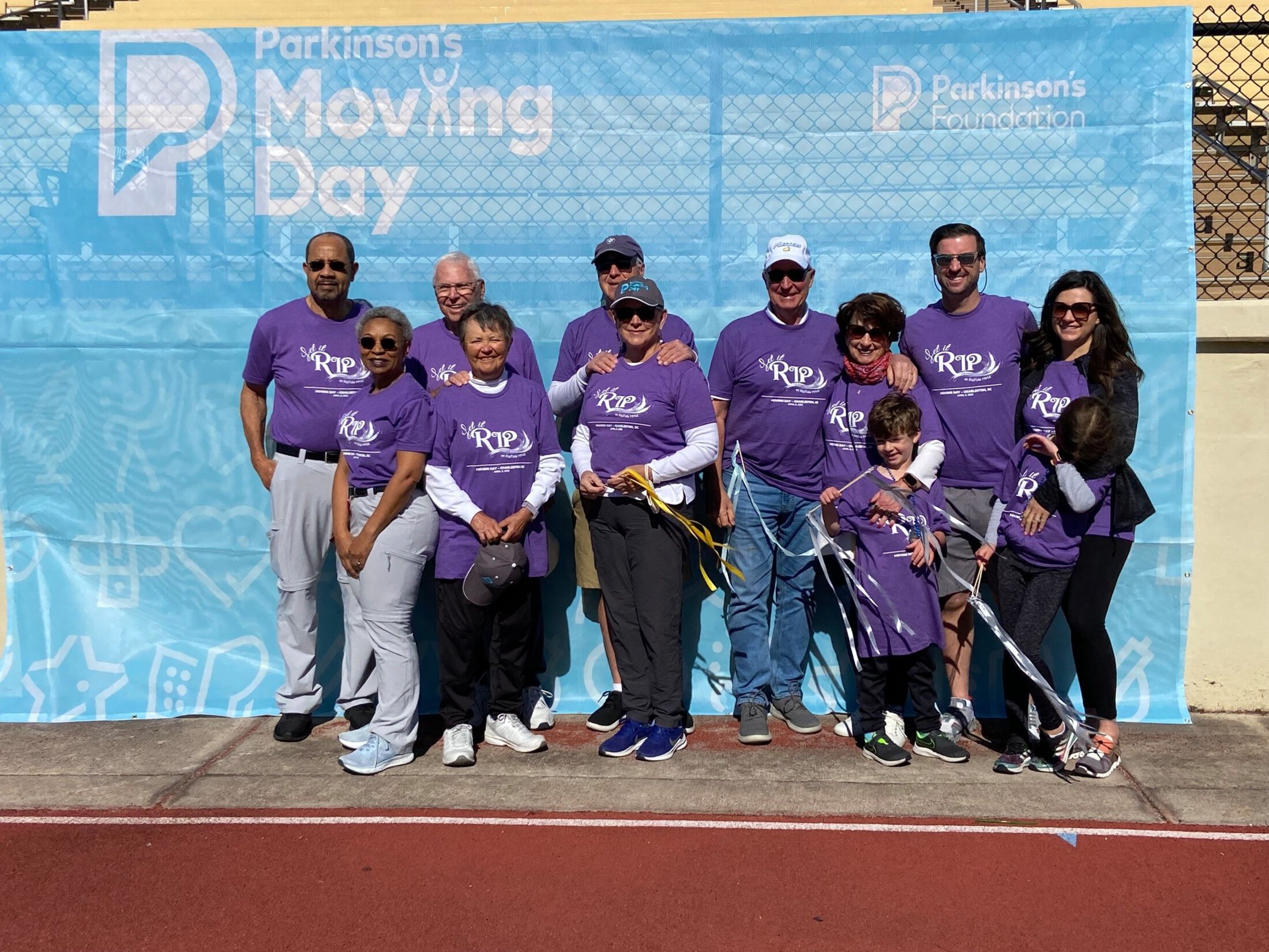A group of people in matching purple shirts stand in front of a Parkinson's Moving Day banner.