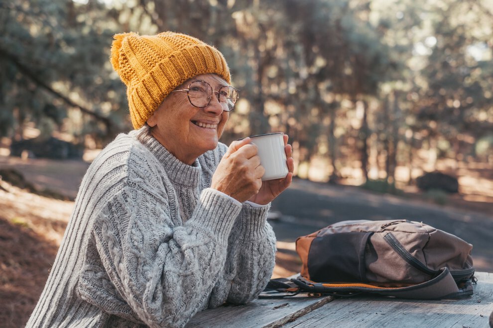 A woman in a thick cable knit sweater and knitted cap drinks coffee from a tin mug while seated at a picnic table in the woods.