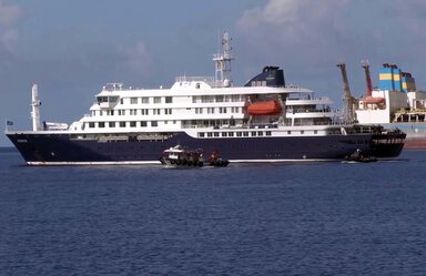 Large blue and white ship on water. It's near a port with cranes for lifting cargo. A red lifeboat is visible.