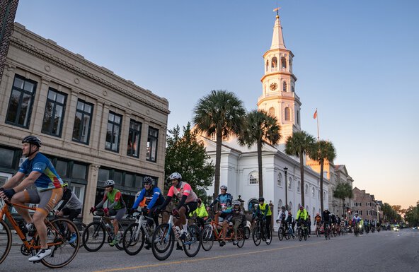 a large group of cyclists rides past a historic church with a tall white steeple in downtown Charleston during the Lowvelo bike ride