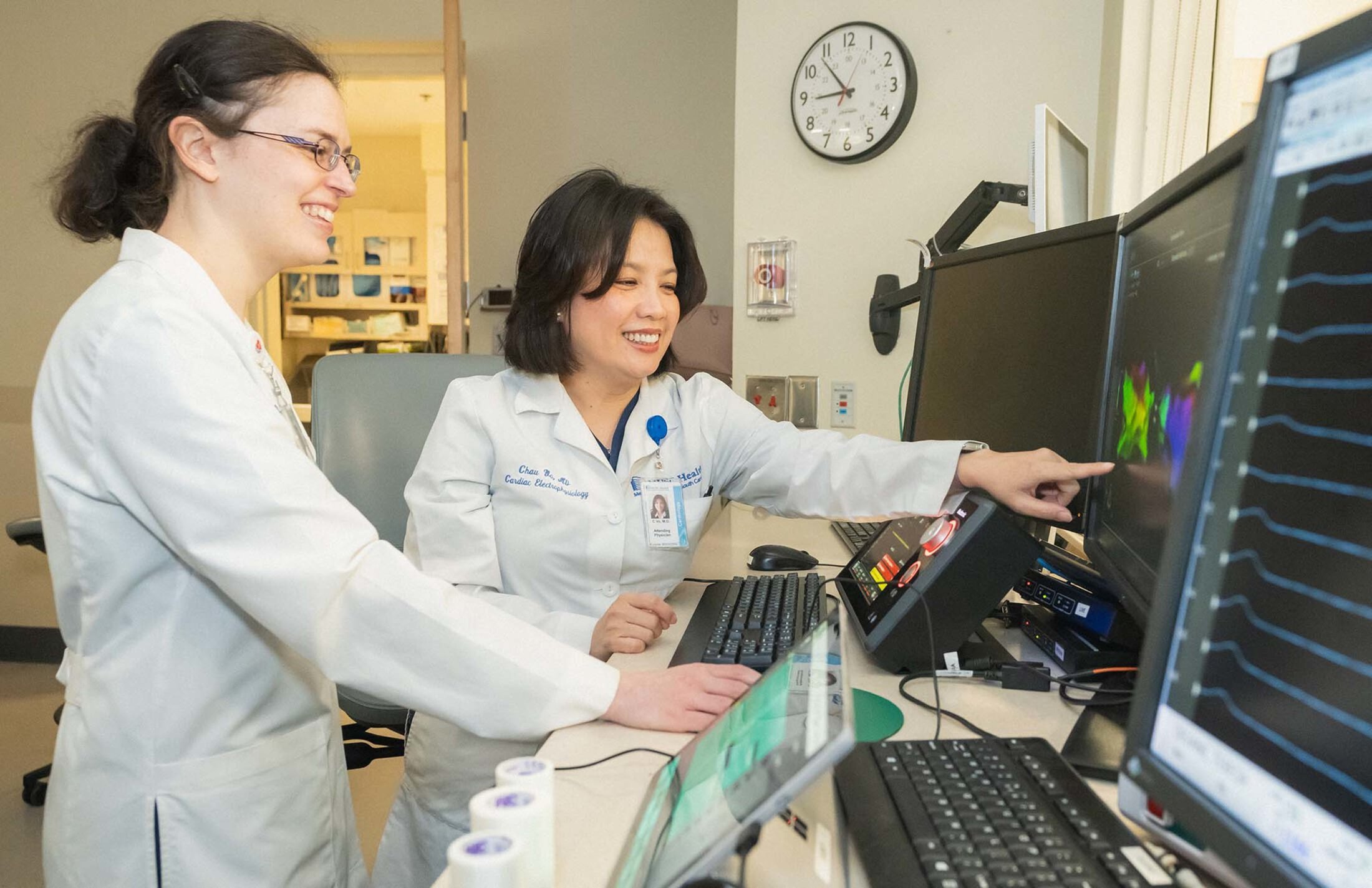 Two women wearing white coats smile as the one on the right points to an image on a computer screen. The one on the left is wearing glasses. There is a clock behind them that says it's about 8:55.