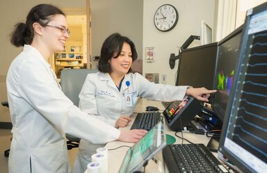 Two women wearing white coats smile as the one on the right points to an image on a computer screen. The one on the left is wearing glasses. There is a clock behind them that says it's about 8:55.