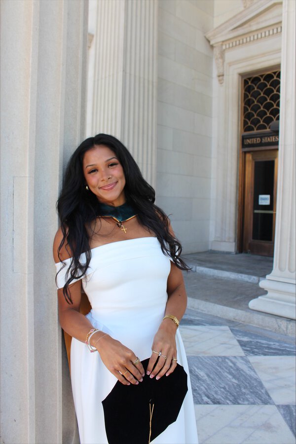 Graduate in a white off-the-shoulder dress poses beside a stone column outside a historic building, smiling while holding a black graduation cap. She wears a green academic hood and gold jewelry, with marble floors and classical architecture in the background.
