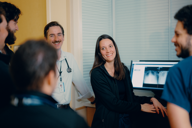 A woman standing with her back to the an image of a chest scan on screen. She smiles at a group of medical professionals who surround her in the room.
