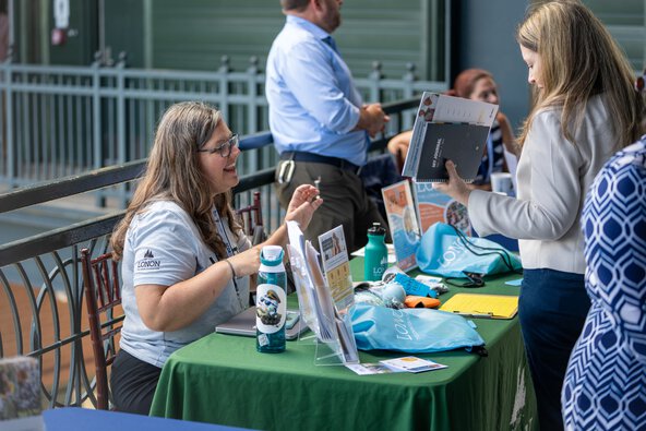 A woman seated behind a table talks with another woman who has stopped to look at the items laid out on the table.