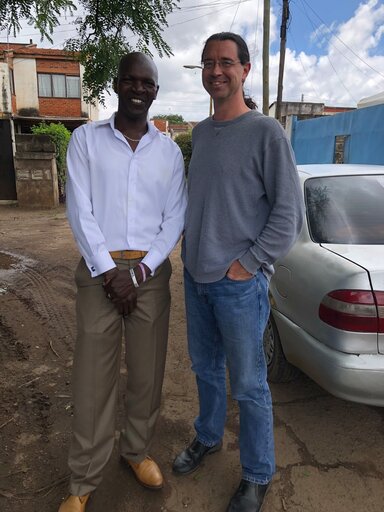 Two men stand in front of a building outside. The man on the left is wearing a white shirt, khakis and a belt. The man on the right has long brown hair in a ponytail and is wearing a gray shirt and jeans. There is a car beside them.