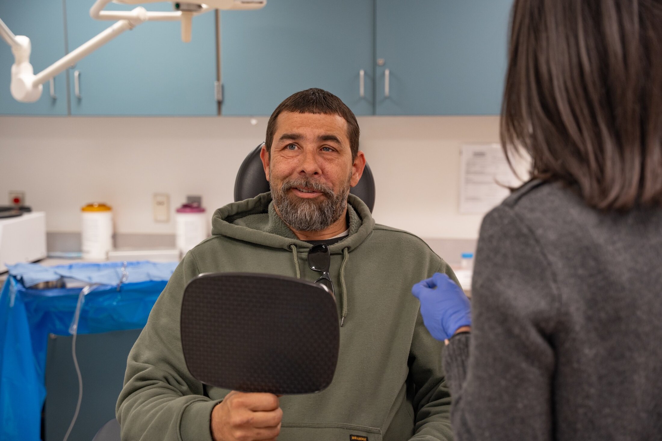 A man smiles looking at someone to his right. He's holding a mirror in his right hand. He's wearing a green hoodie and has dark hair and a short beard.