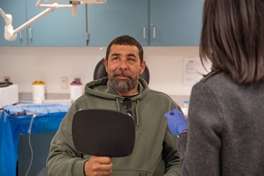 A man smiles looking at someone to his right. He's holding a mirror in his right hand. He's wearing a green hoodie and has dark hair and a short beard.
