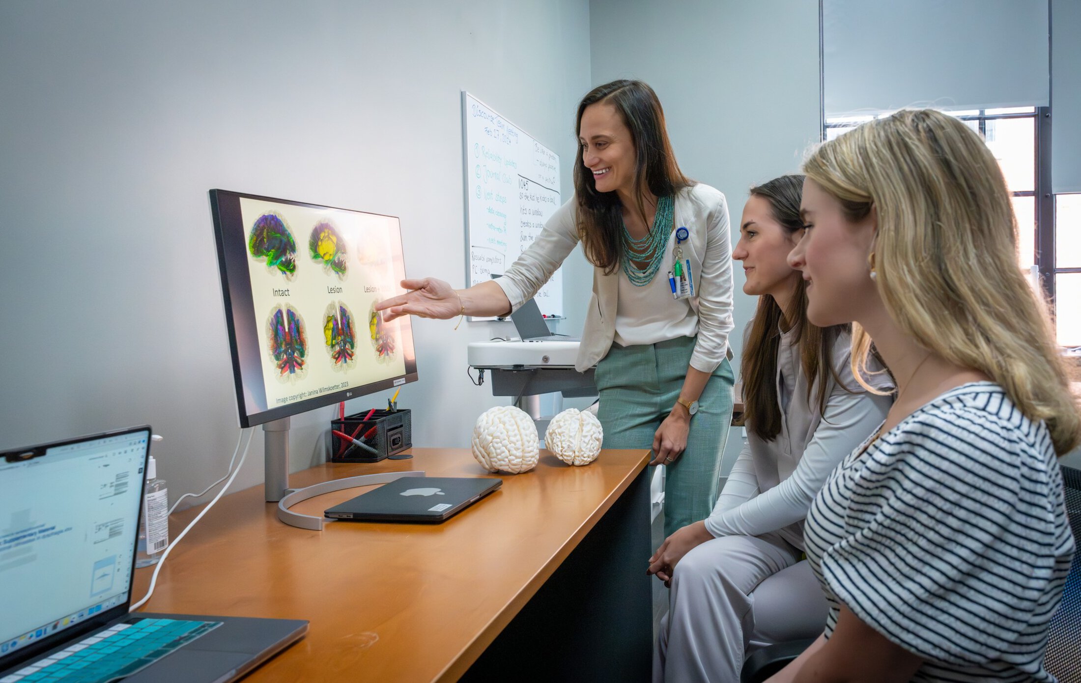 A woman stands in front of a desktop screen and points to the image. Two seated female students observe.