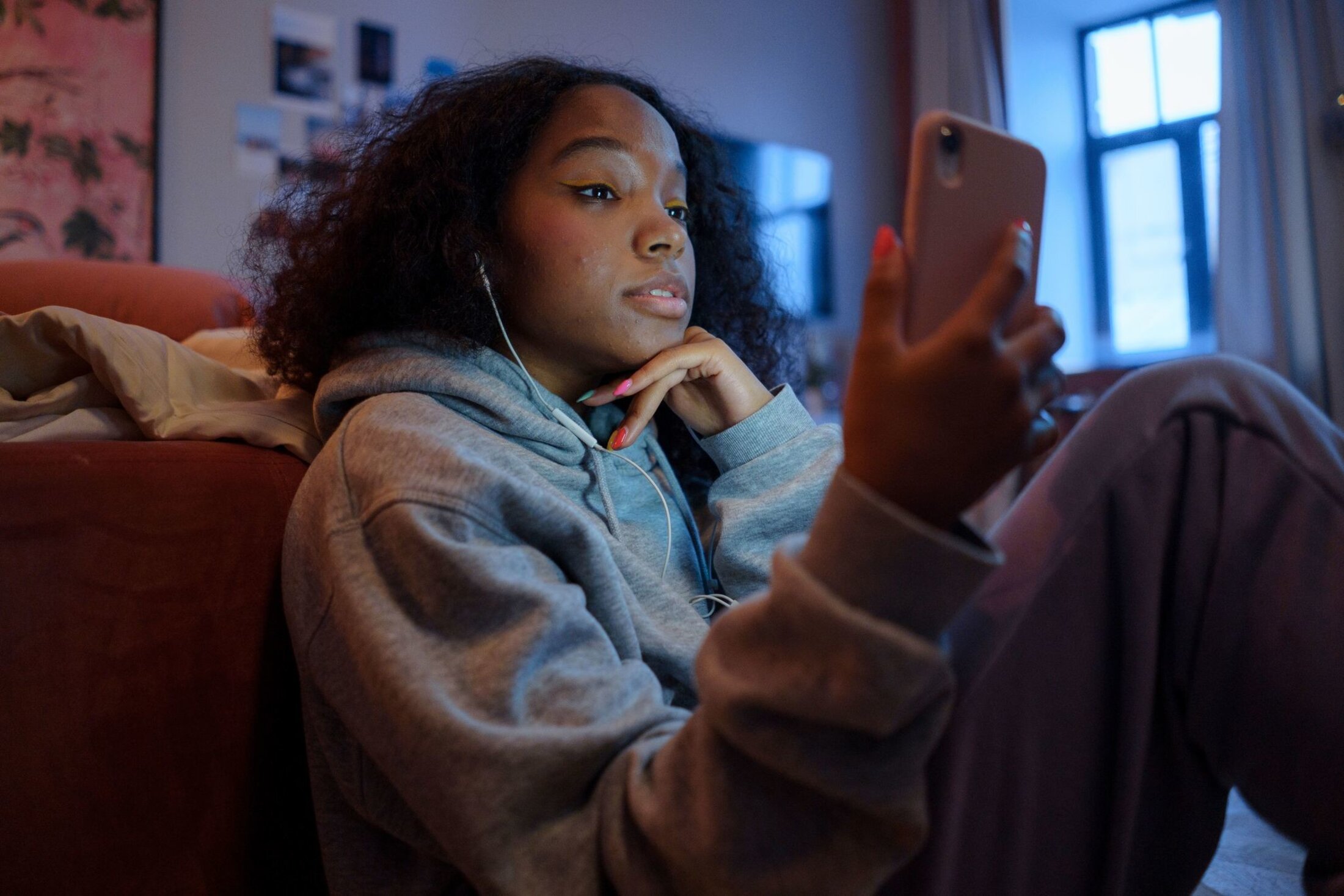 Young woman is sitting and looking at a pink covered cell phone. She's wearing a gray hoodie and sweatpants. Her left hand is under her chin. She has long, multi-colored fingernails.