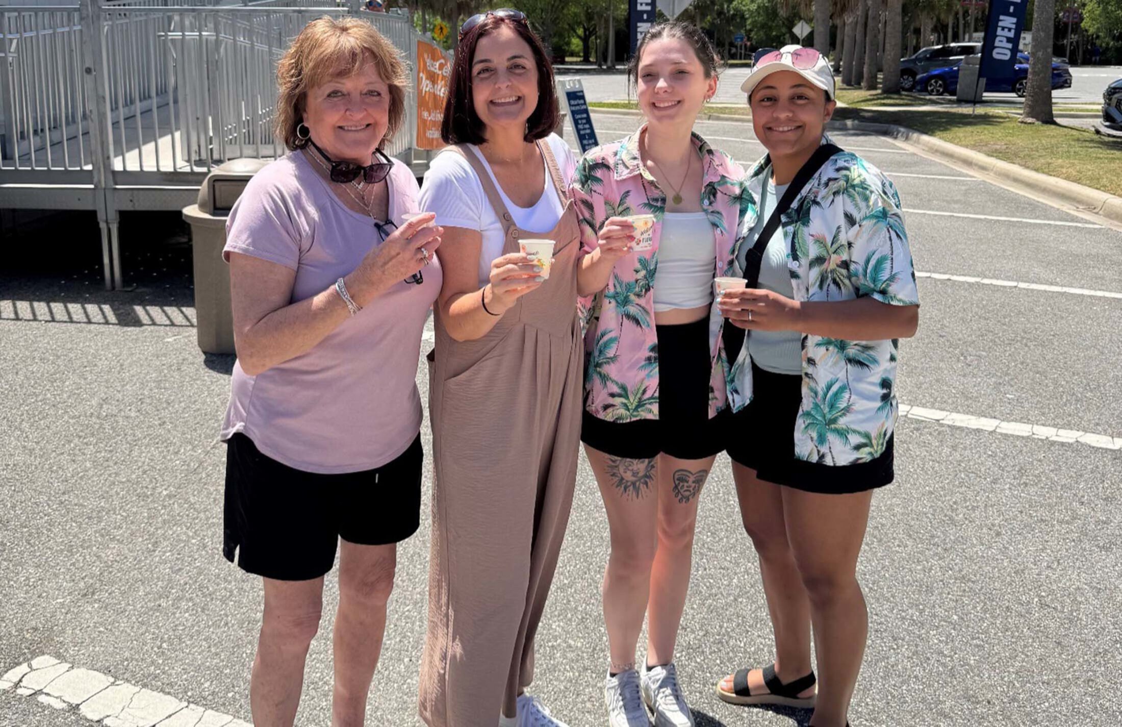 Four women stand outside holding cups. The woman on the far left is wearing a pink t-shirt and black shorts. Next to her is a woman wearing overalls and a t-shirt. The two women on teh right are wearing Hawaiian shirts and black shorts.