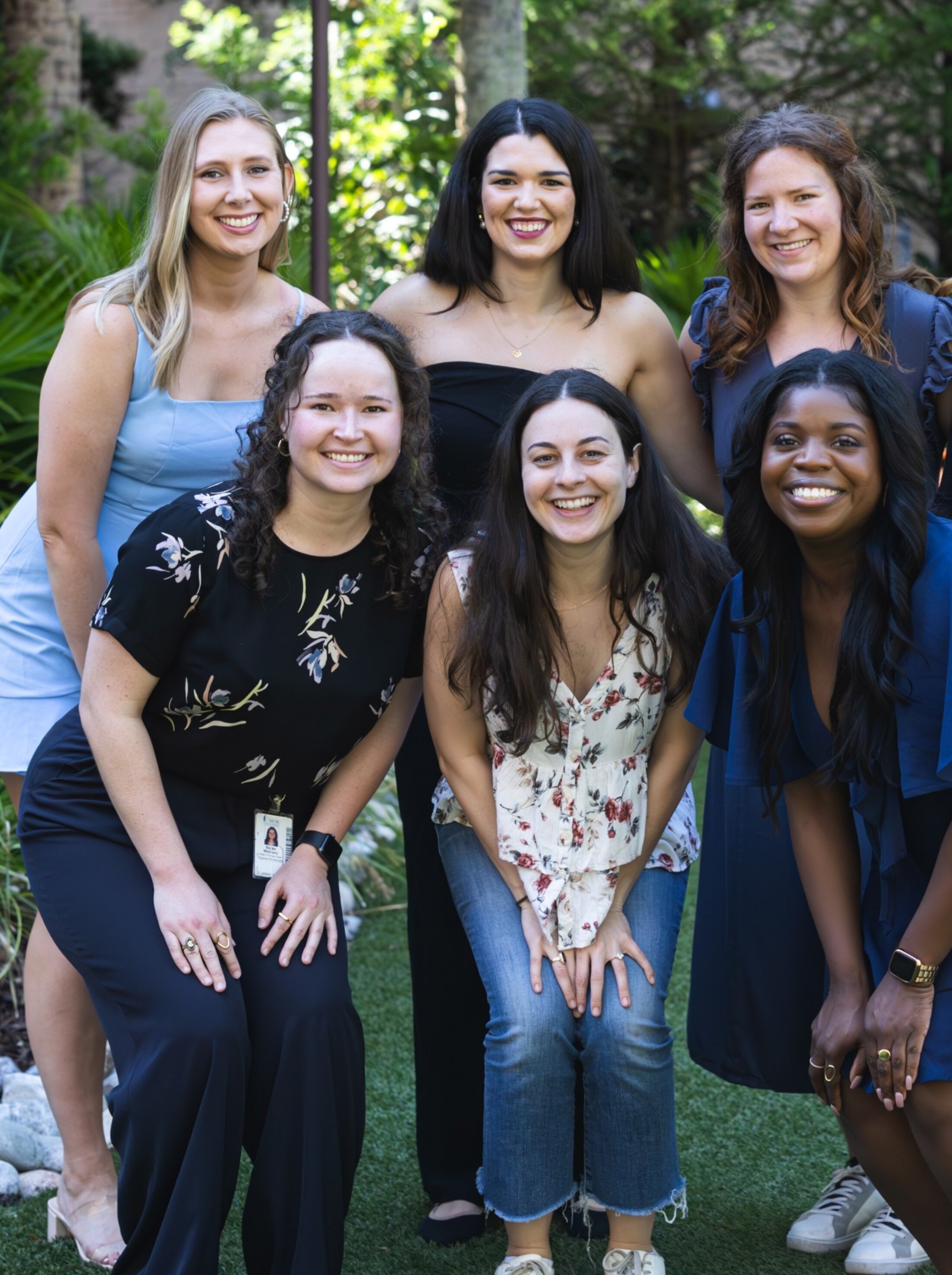 Six women pose together outdoors, smiling at the camera. They are standing on grass with greenery and trees in the background.