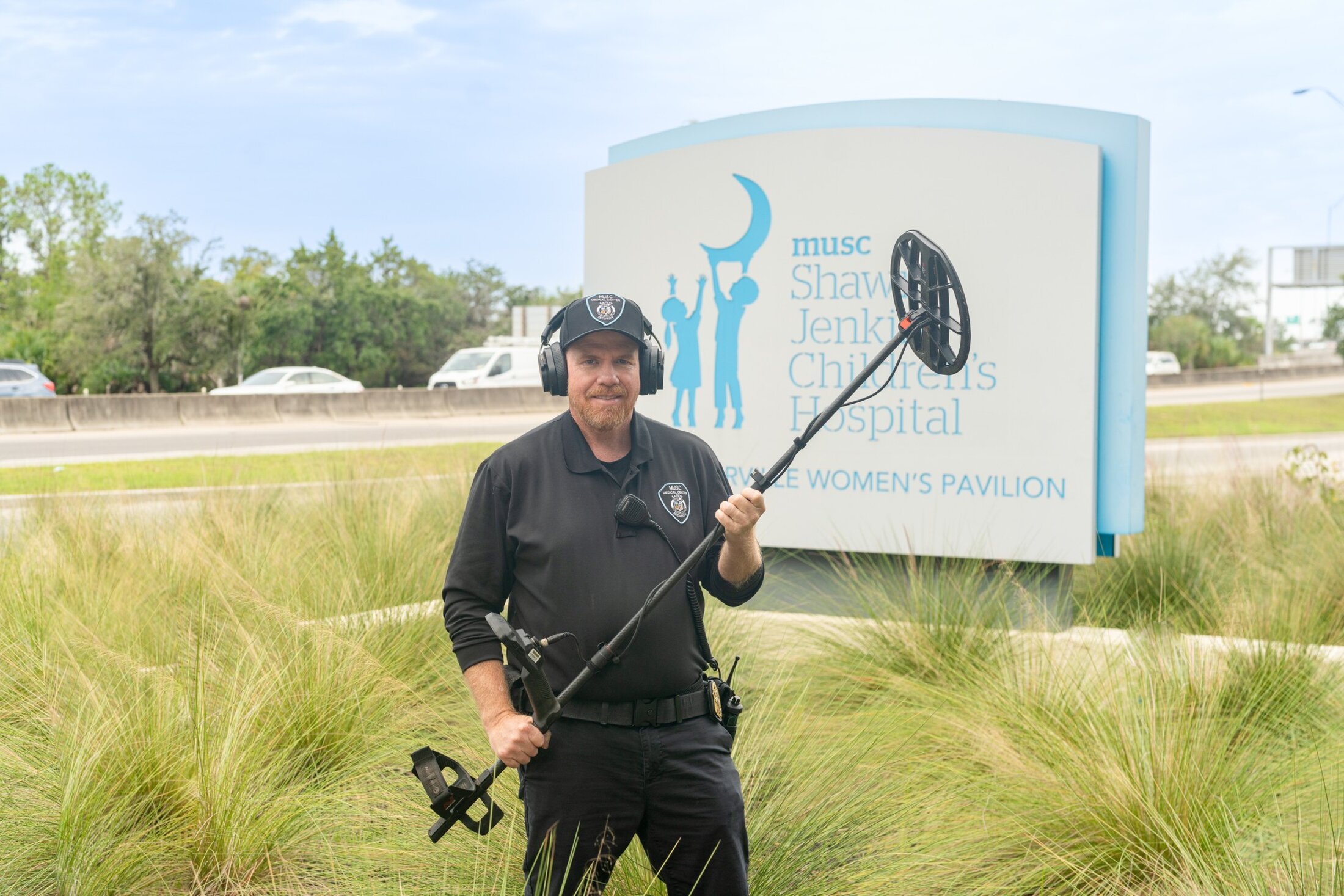 A man wearing a black polo shirt and ballcap with the MUSC security logo stands by the MUSC Shawn Jenkins Children's Hospital and Pearl Tourville Women's Pavilion sign.