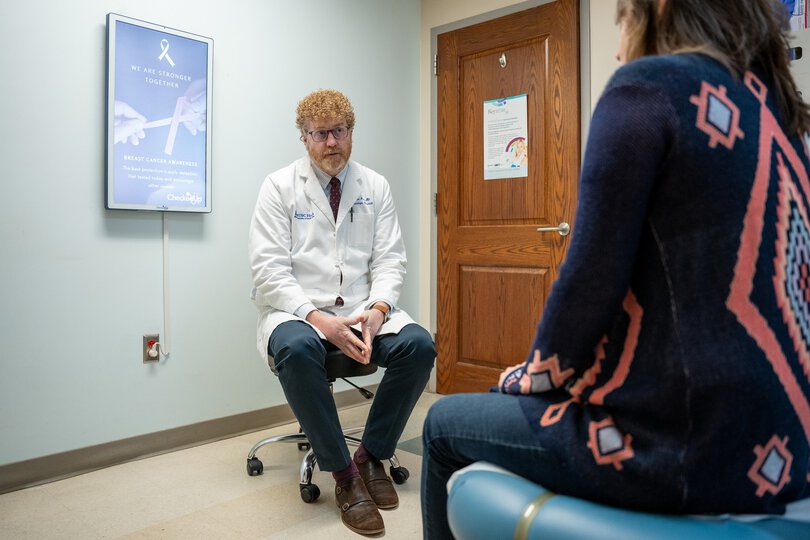 A doctor sits on a stool in an exam room and speaks with a patient.