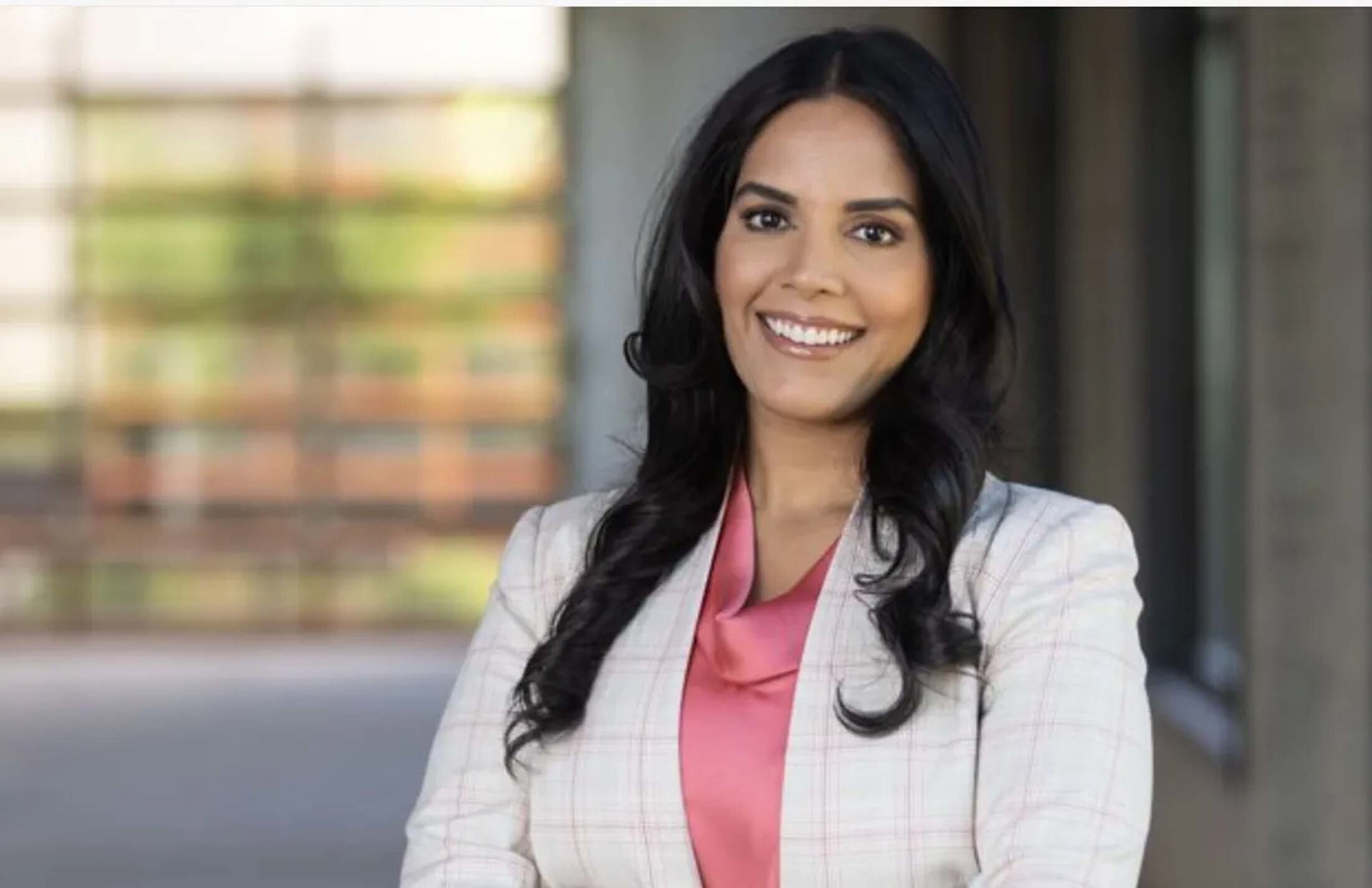 Woman with long dark hair smiles. She is wearing a pink shirt under a grey jacket. The background is blurred.