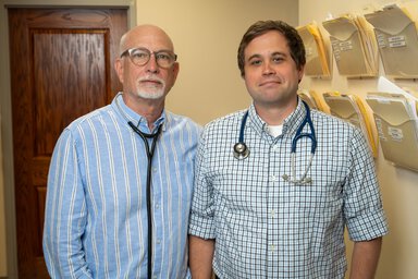 Two doctors in button-down shirts open at the collar & stethoscopes around their necks pose in a clinic hall