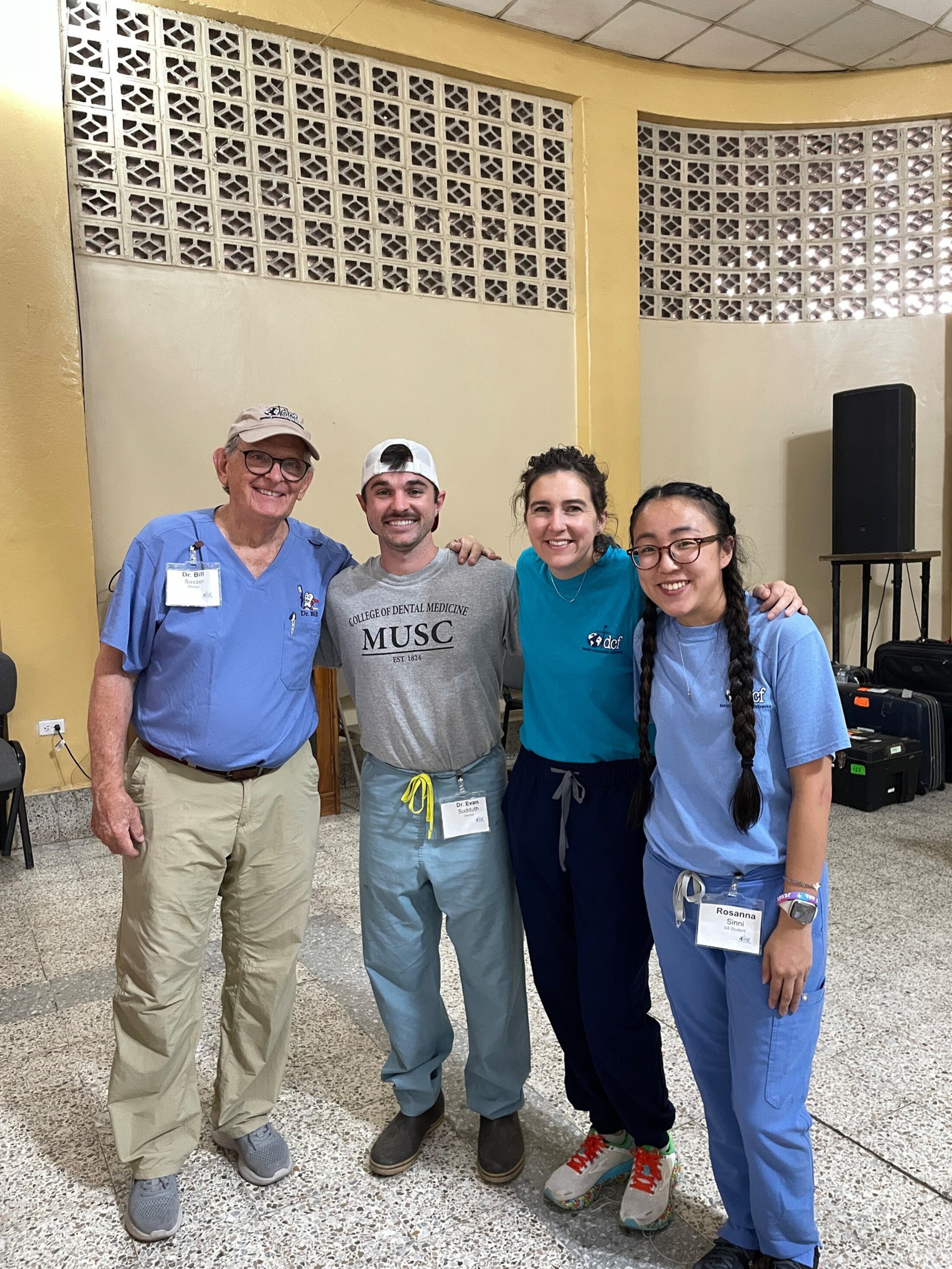 our people standing together and smiling inside a large room with light-colored walls and decorative block windows. Two people wear medical scrubs and name badges, and the group poses with their arms around one another during what appears to be a healthcare or volunteer event.