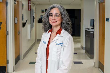 A doctor in a white lab coat poses in a medical office hallway.