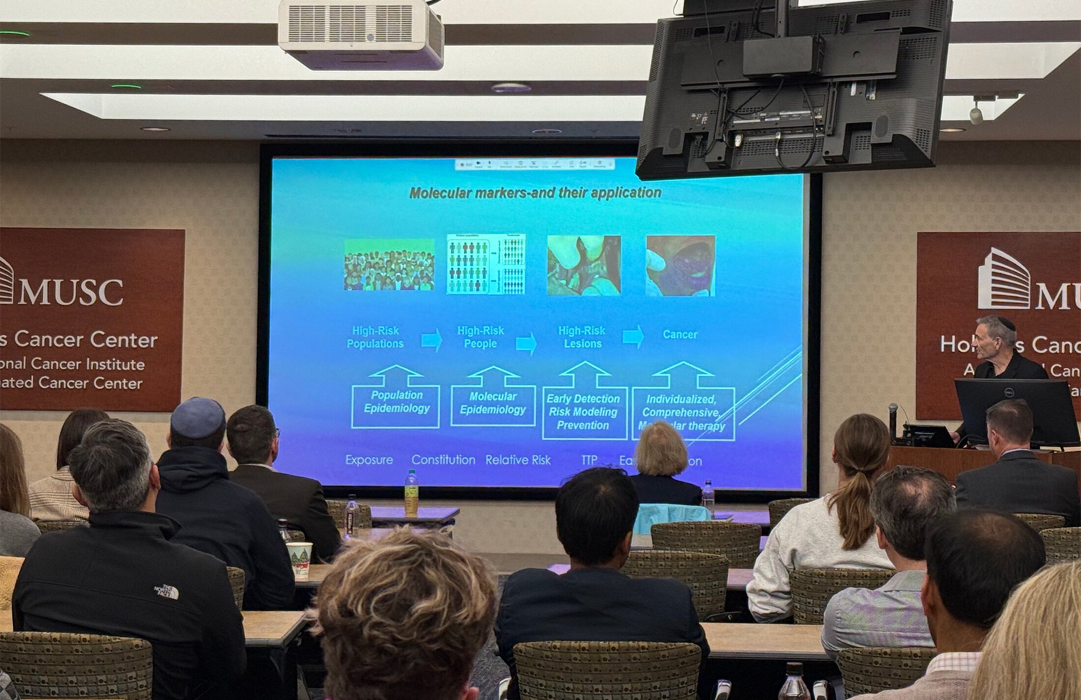 An audience looks at a slide on a large screen while a man speaks at a podium.