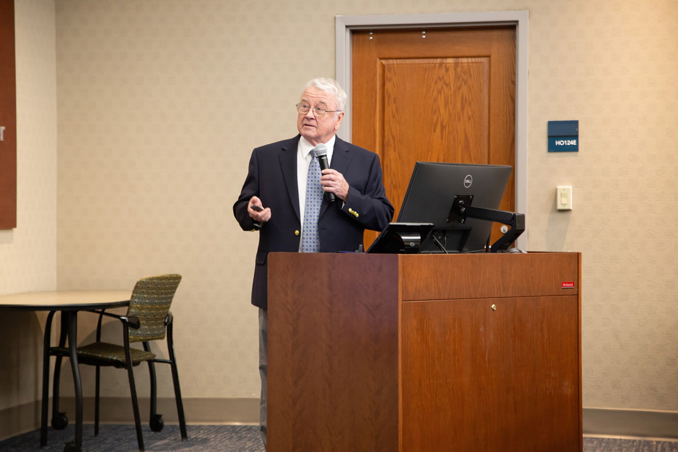 A man speaking at a podium gestures to the screen showing his slides.