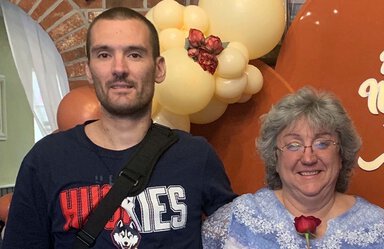 A man and a woman stand in front of yellow balloons. The man, younger than the woman, has dark hair and is wearing a T-shirt that says Huskies. The woman has gray hair feathered back, eye glasses, a blue top and is holding a red rose.