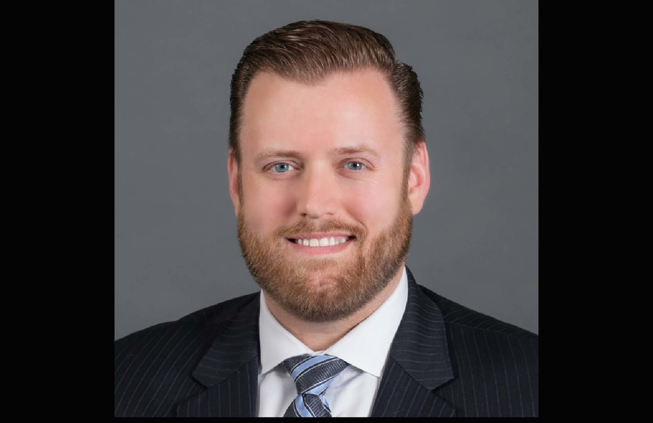closeup of man with light brown hair and a beard. He is smiling and appears to be wearing business clothing.