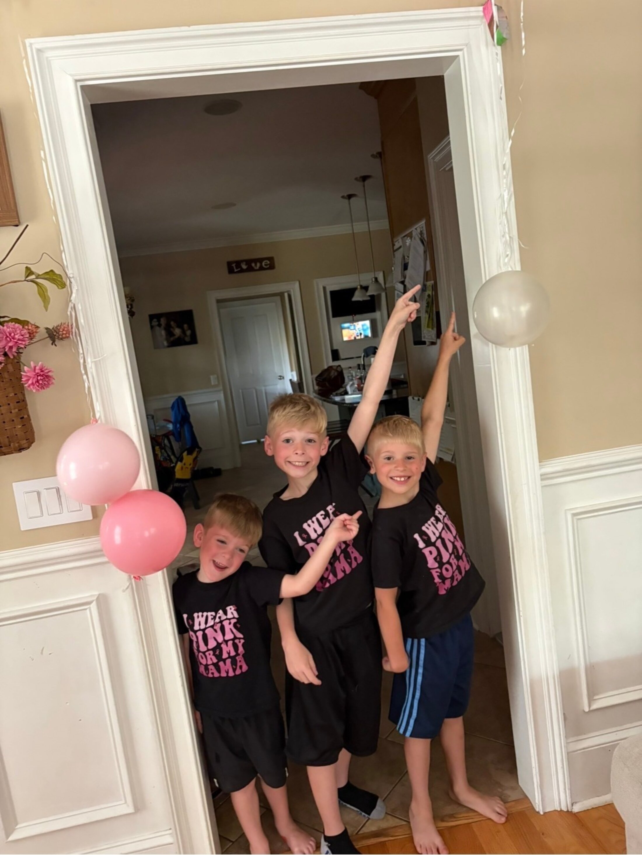 Three little boys, obviously brothers, all wear shirts that say 'I wear pink for my mama' and point upwards at a balloon in their home.