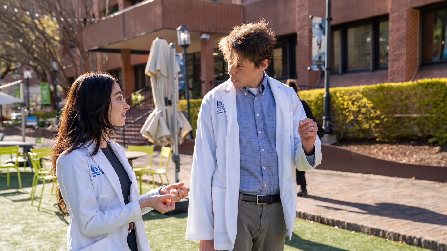 Two young people in medical white coats chat as they walk along a pedestrian road.