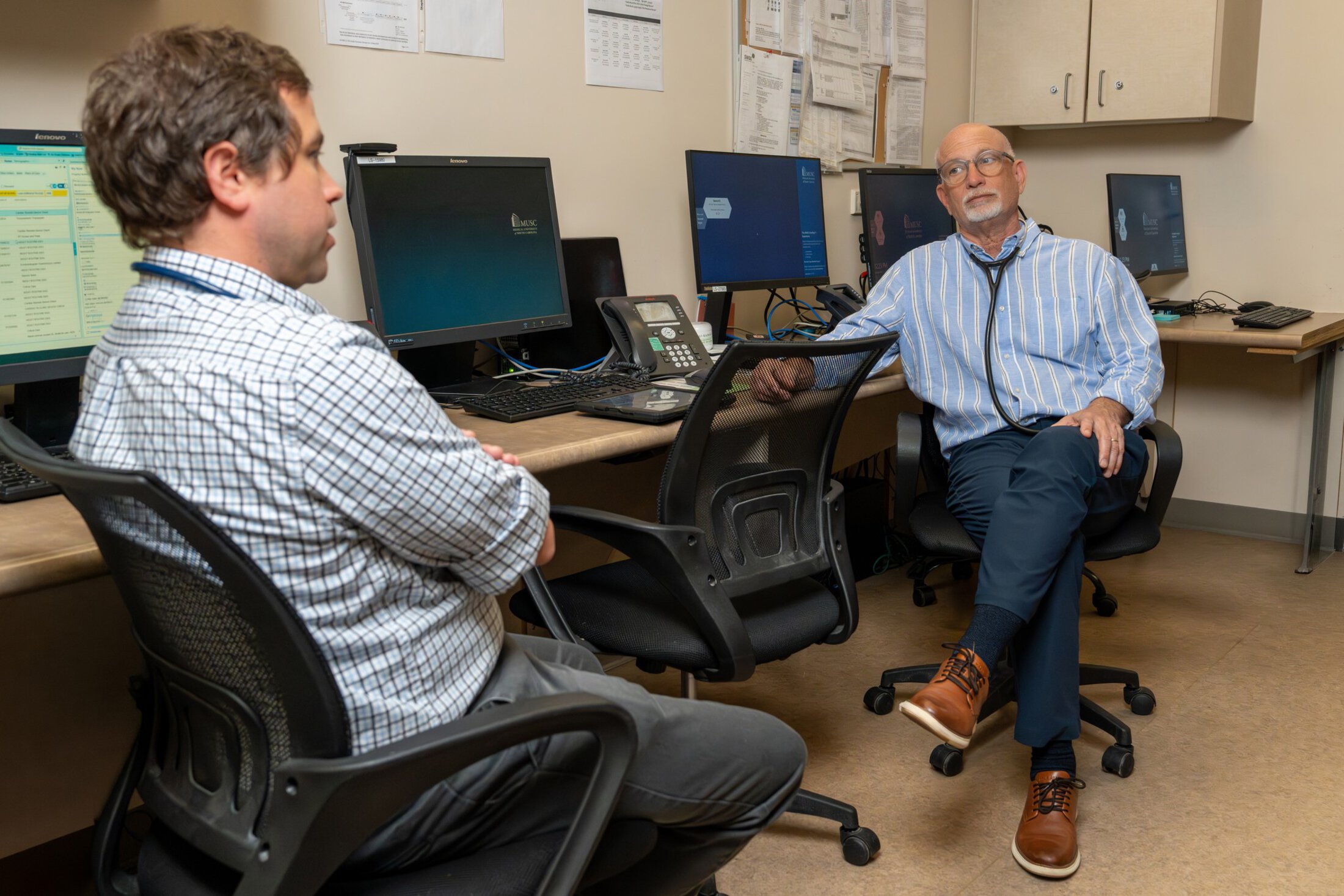Two doctors sit at a table lined with computers and talk.