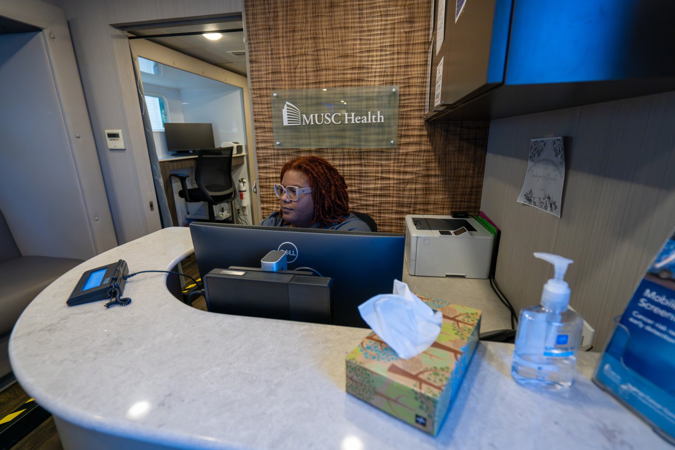 A receptionist sits behind a computer screen with a door to the clinic area behind her.