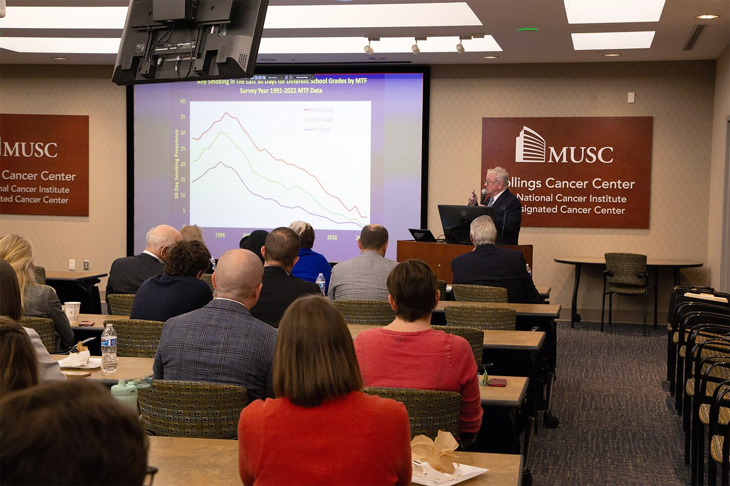 A man speaks at a podium with a large screen showing slides behind him.