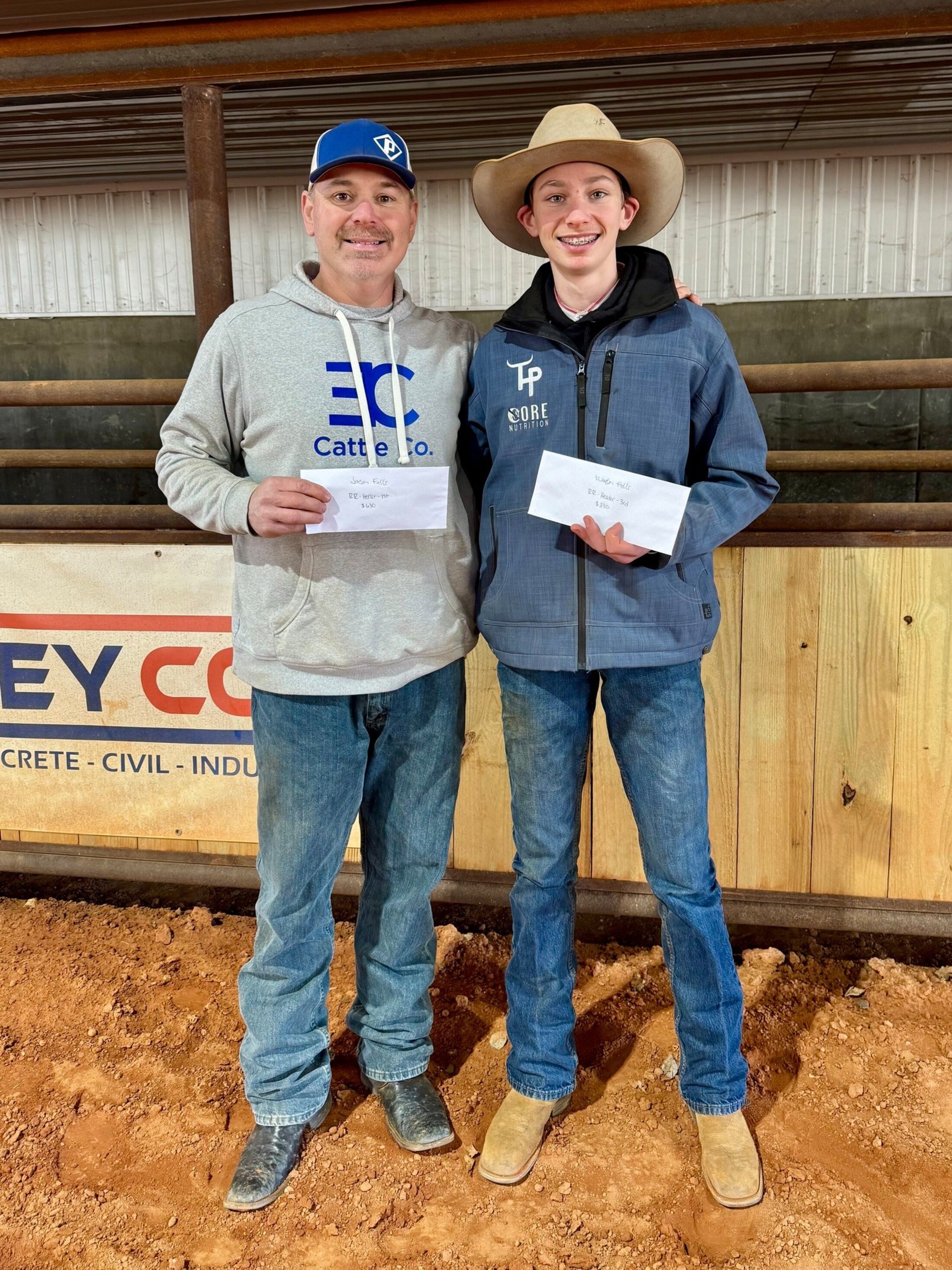 A middle-aged man in jeans, sweatshirt and baseball cap poses with a teen boy in jeans, jacket and cowboy hat in a rodeo arena, holding envelopes containing their winnings.