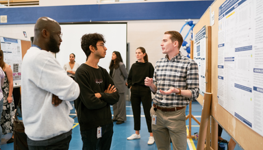 Male student stands in front of his poster talking to other male students.