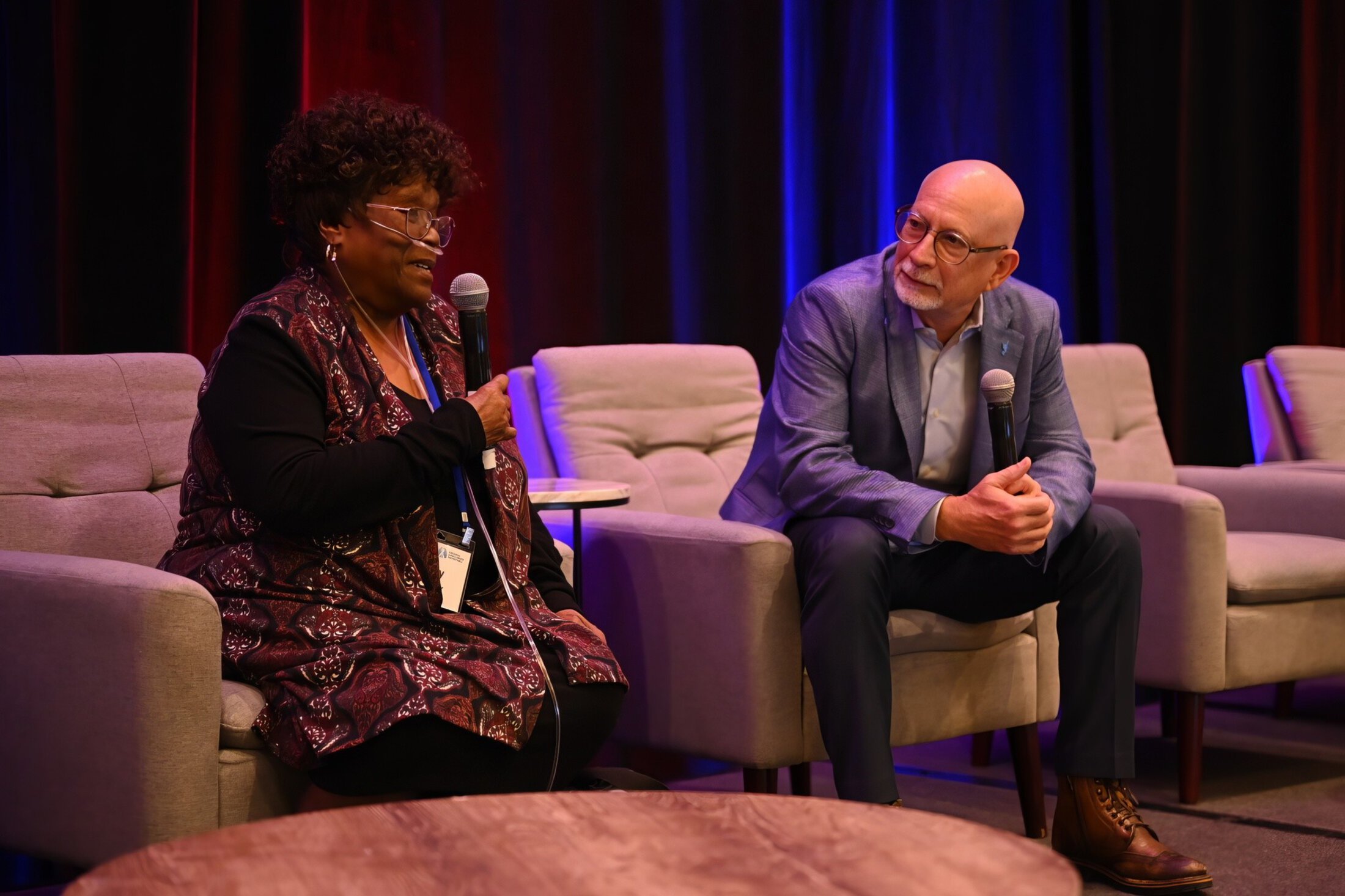 Two people sit in recliners on stage with microphones, the man leaning forward, elbows resting on knees, to listen to what the woman is saying.
