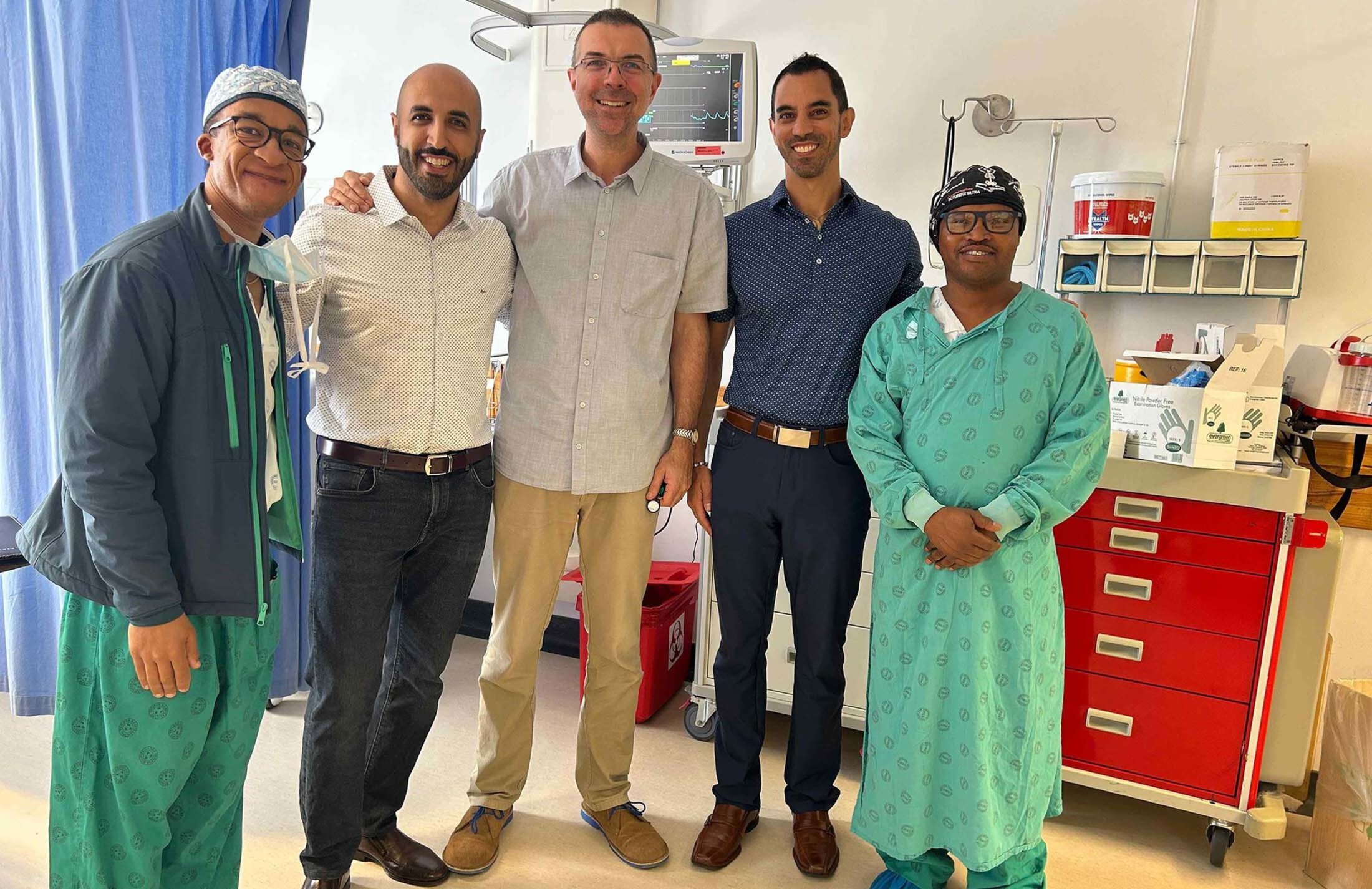 Five men stand together smiling. The men on both ends of the group are wearing hospital scrubs. Behind the group is a medical screen and cabinet.
