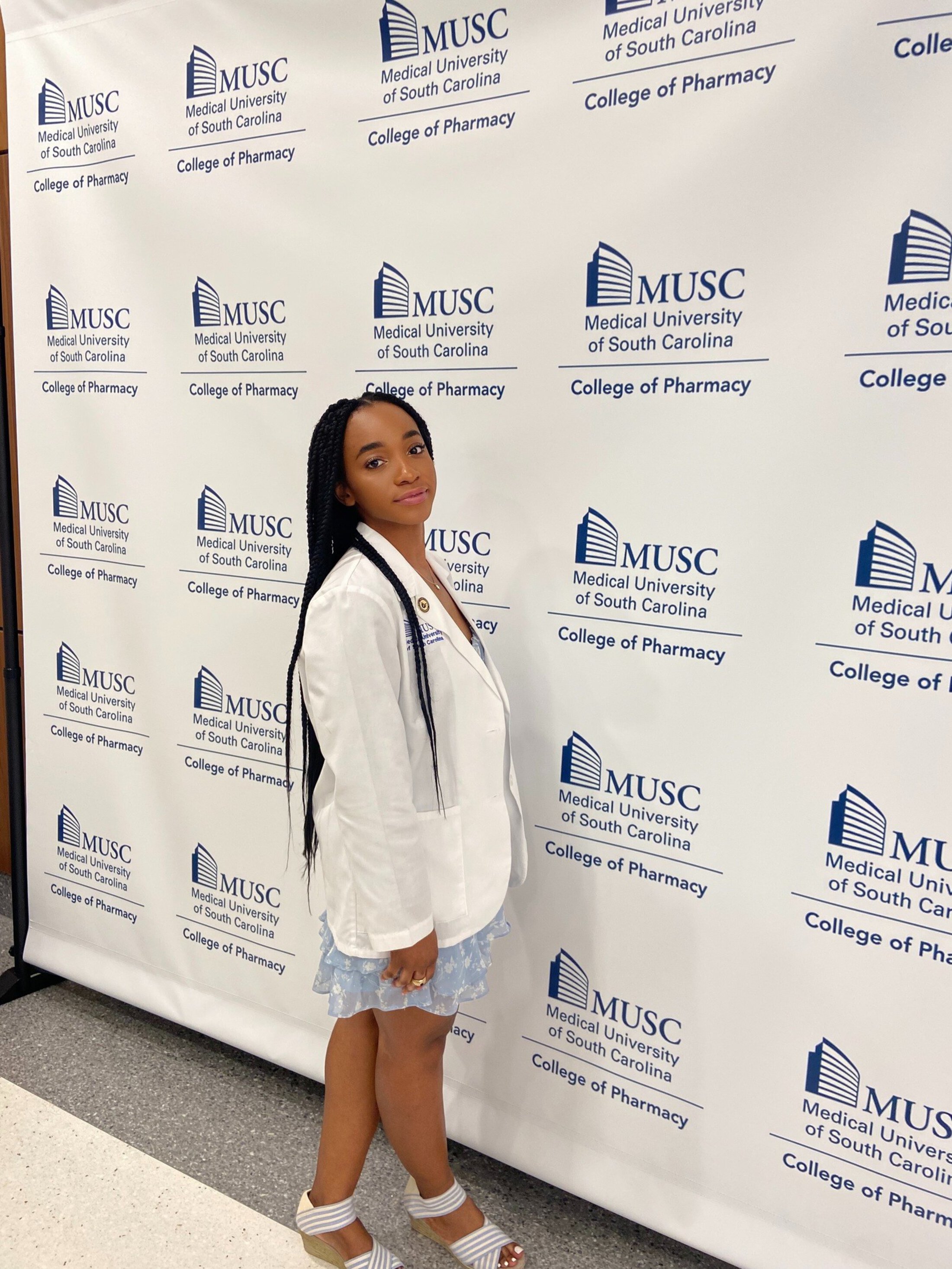 Woman in a white coat stands in front of a college of pharmacy backdrop.