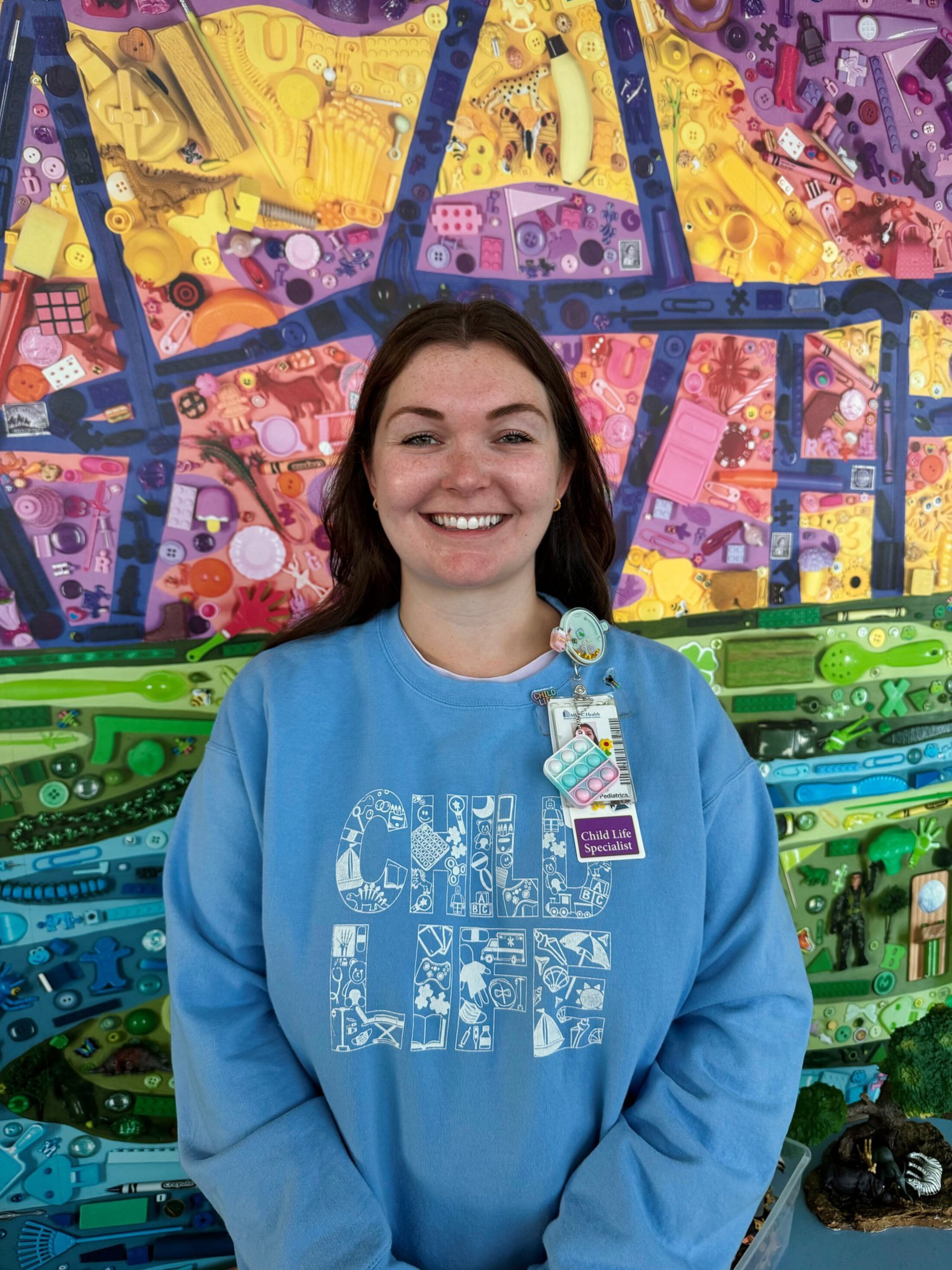 Smiling woman wearing a light blue “Child Life” sweatshirt and a hospital badge labeled “Child Life Specialist,” standing in front of a colorful wall mural made of small toys and objects arranged in rainbow sections.