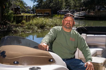 A man poses in the captain's chair of a pontoon boat docked on a creek that winds between back yards.