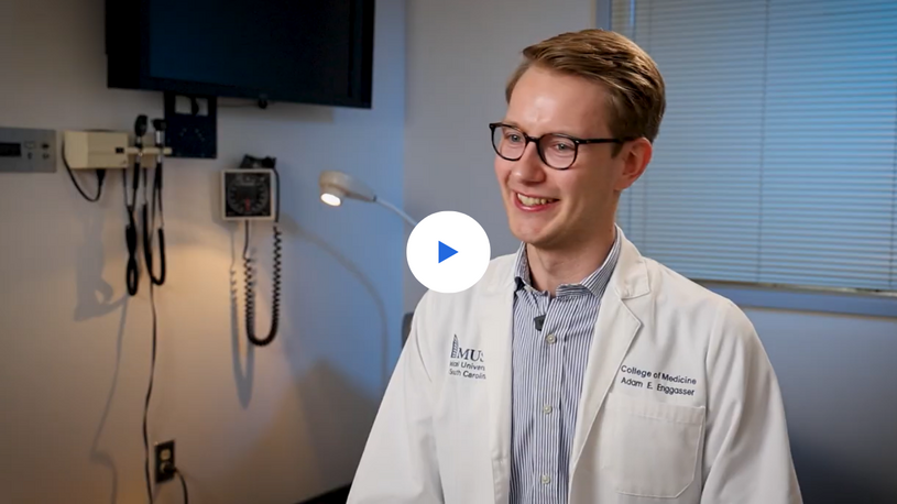 Smiling male medical student in glasses and white lab coat sits in a clinical exam room, with medical equipment behind him and a play icon.