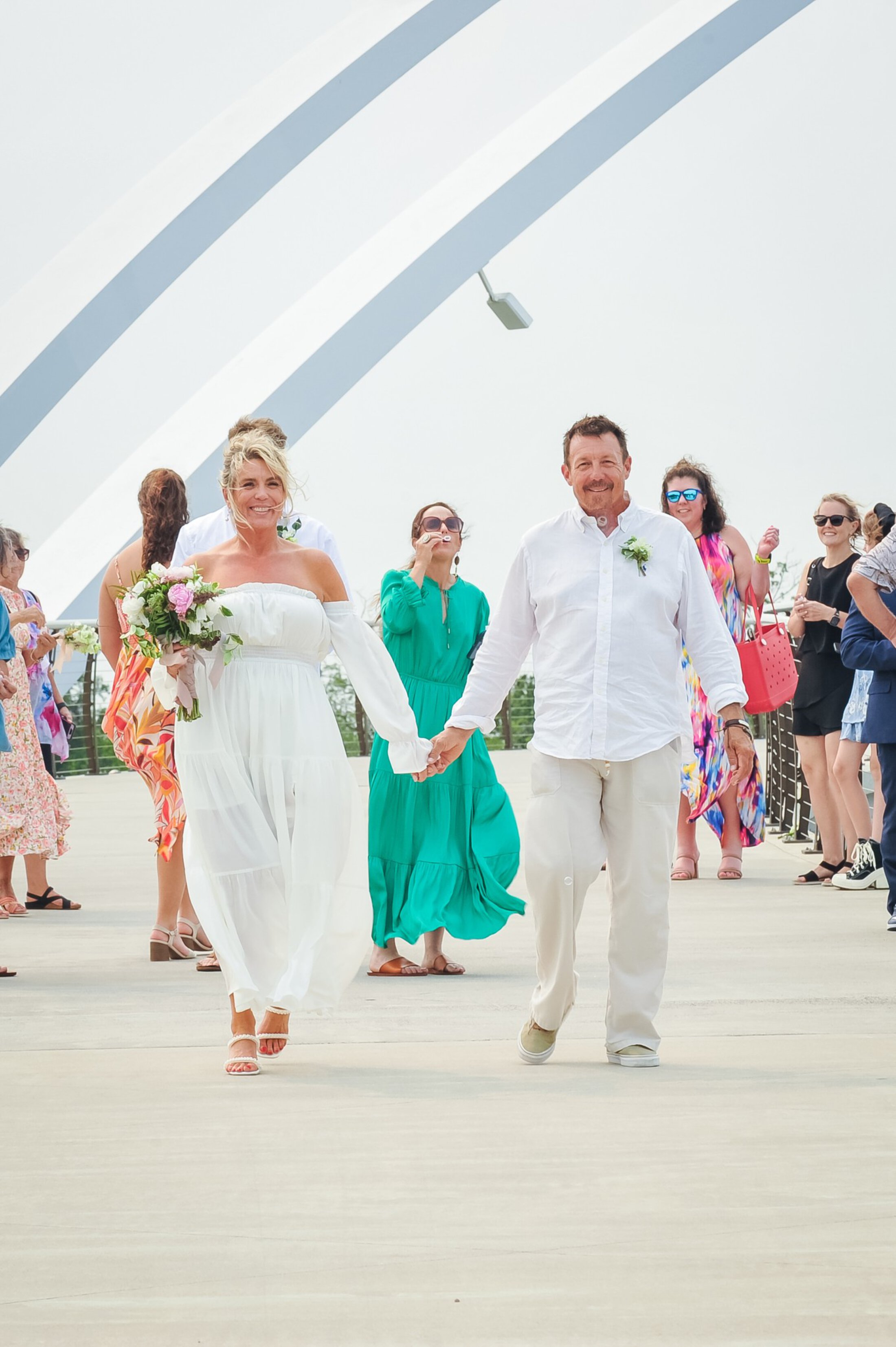 A coastal-casual bride and grown beam as they walk hand in hand and a woman behind them blows bubbles.