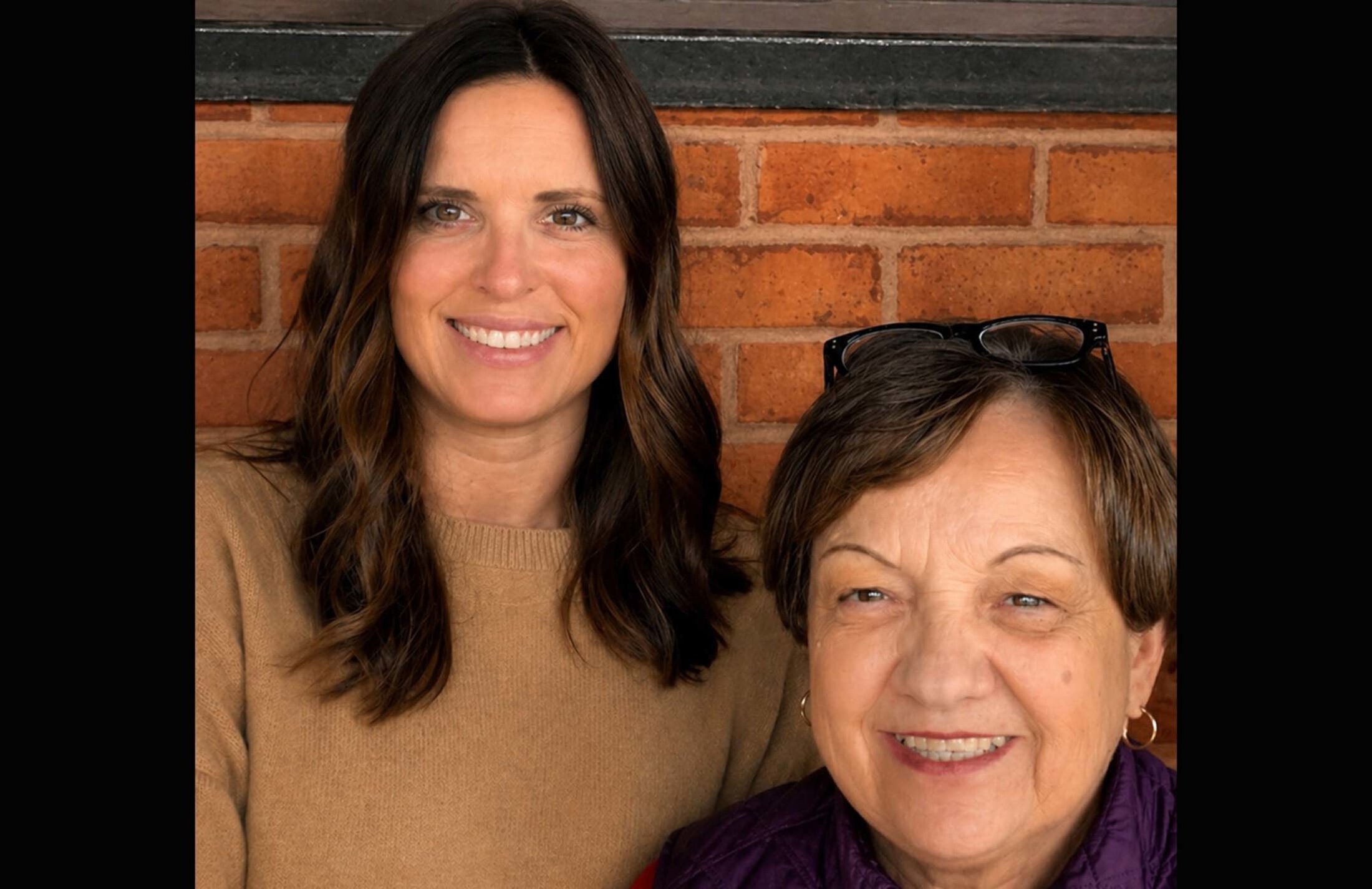 Two women smile. The one on the left is taller. She has long brown hair and is wearing a tan sweater. The one on teh right has short brown hair and is wearing a purple top.