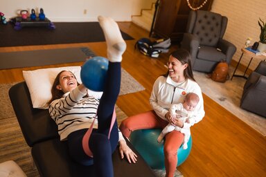 A woman sits on a stability ball while holding an infant and observing a patient performing a pelvic health rehabilitation exercise on a treatment table.