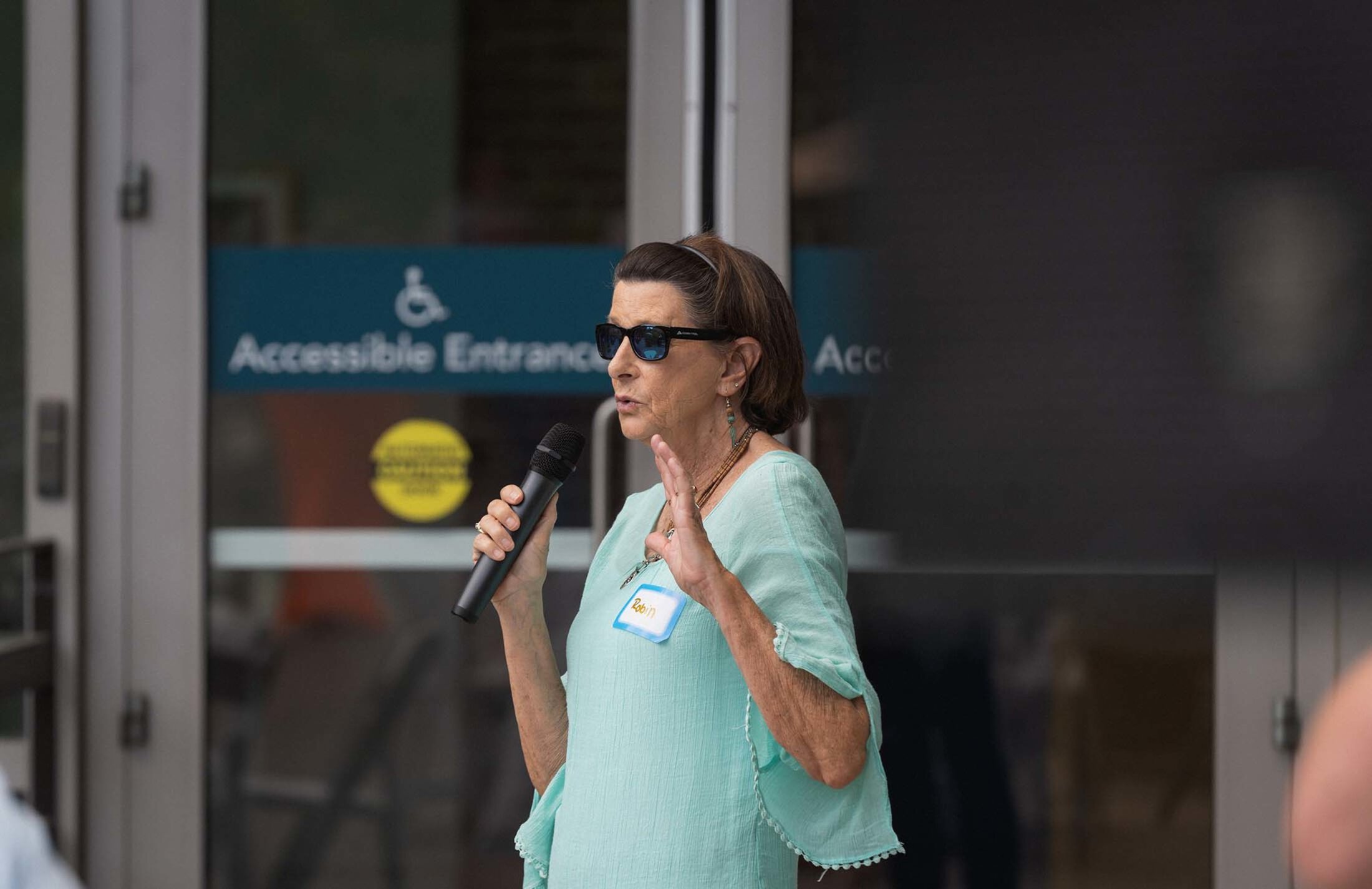A woman wearing sunglasses, a turquoise top and a hairband holds a microphone as she speaks. She is in front of a door that has a picture of a wheelchair and the words accessible entrance.