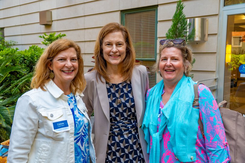 three women pose and smile at an outdoor event in a garden.
