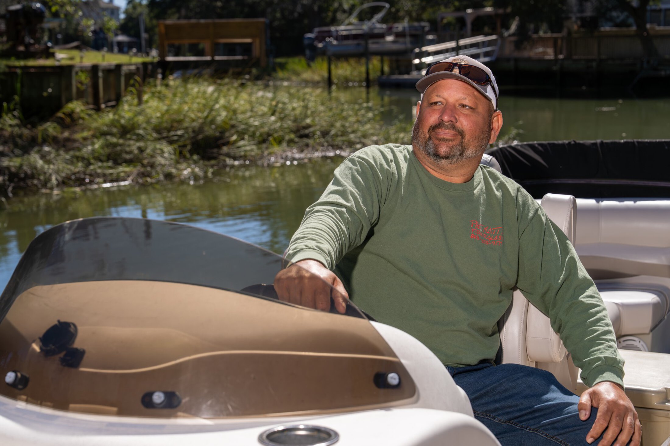 a tanned, robust-looking man sits in a pontoon boat on a creek, gazing into the distance
