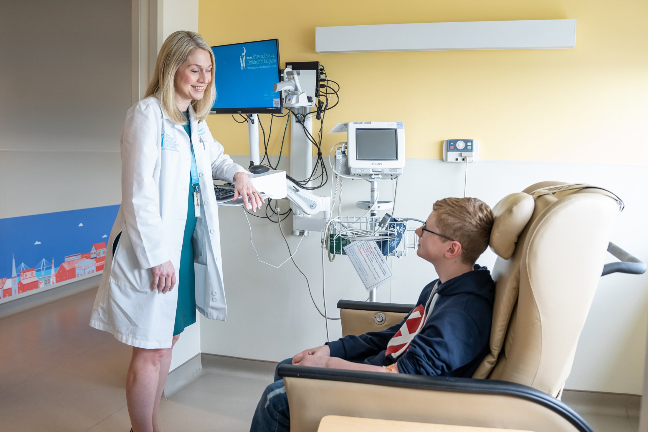 A doctor leans against the computer shelf as she talks to a young patient in a recliner.
